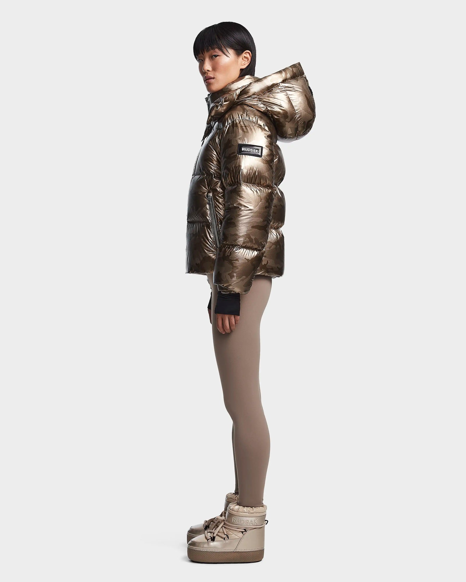 A person stands in profile against a plain white background wearing the Rudsak MAVE S - 8123563 Metallic Ivory Camo water-repellent jacket with lightweight insulation, styled with taupe leggings and platform winter boots.