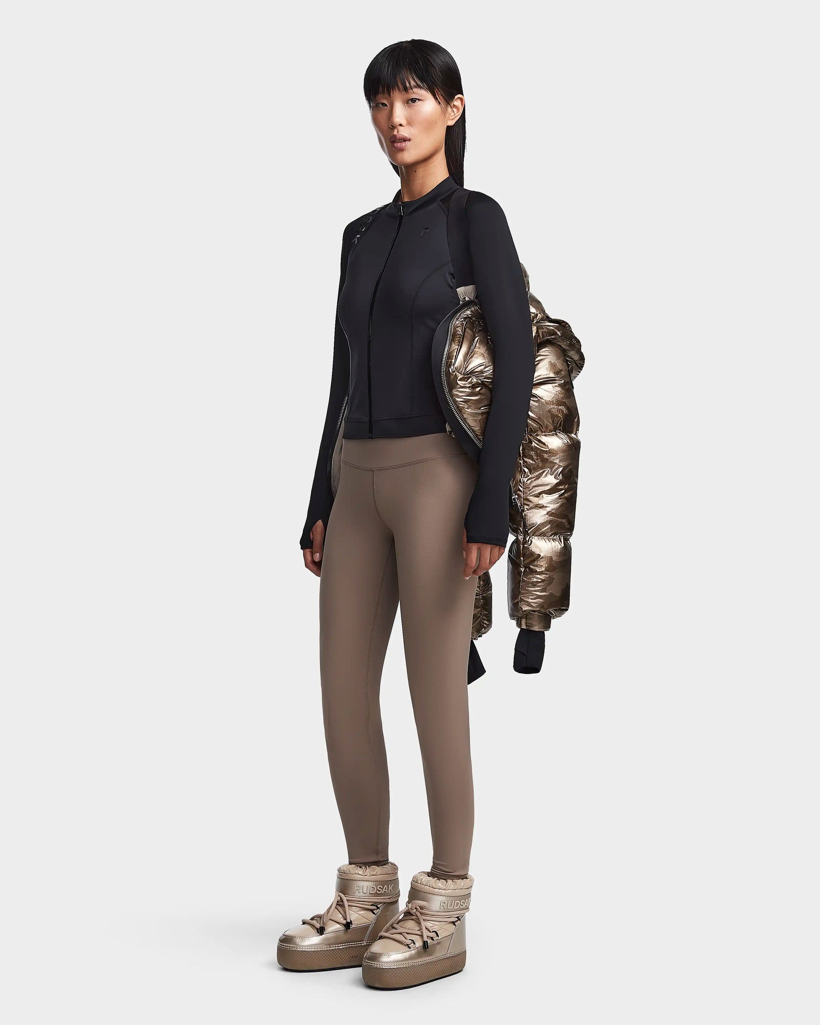 A woman with straight, dark hair and bangs stands facing slightly left in a fitted black jacket, taupe leggings, and metallic gold boots. She holds the Rudsak MAVE S - 8123563 Metallic Ivory Camo water-repellent jacket draped over one arm.
