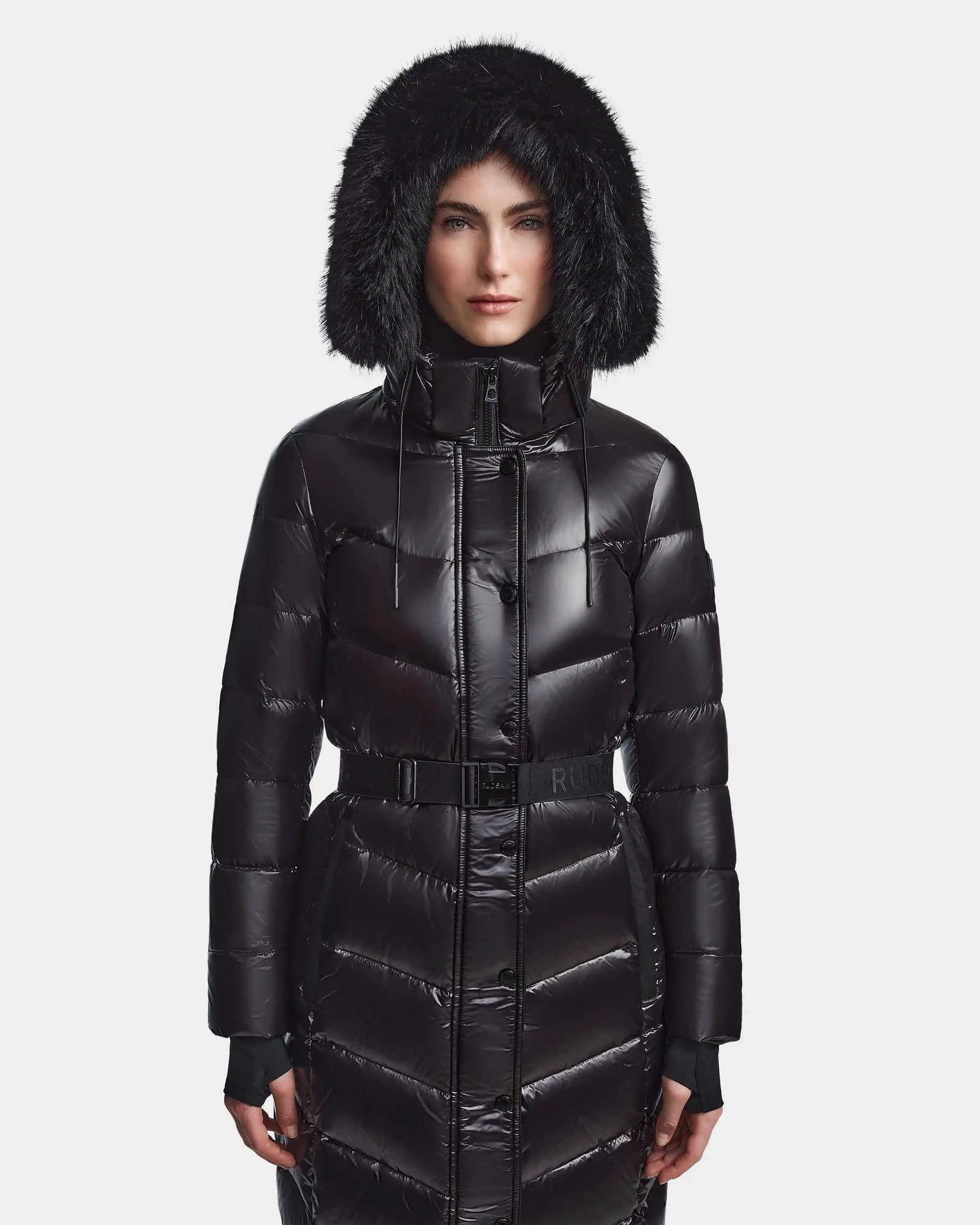 A person wears the Rudsak ELSIE S - L123518 Black women’s maxi down puffer, featuring a shiny finish, belted waist, and a hood with black faux fur trim. They face forward against a plain white background with the hood up.