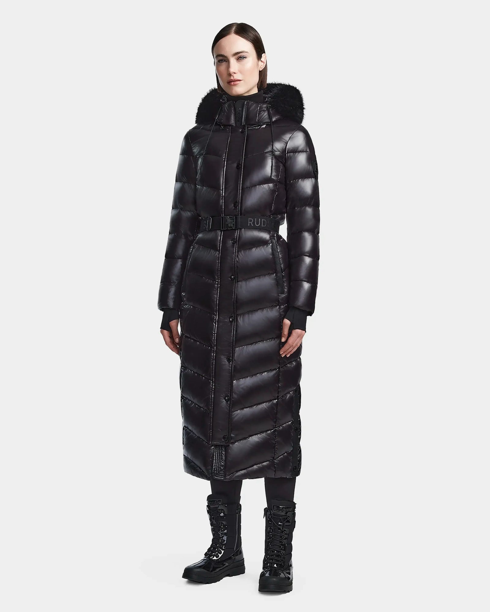 A woman stands against a white background wearing the Rudsak ELSIE S - L123518 Black maxi down puffer with a belted waist and fur-trimmed hood, styled with black leggings and lace-up boots.