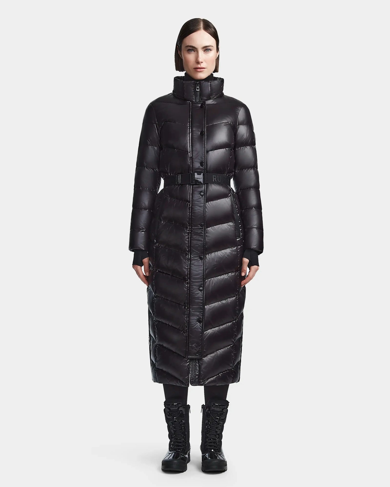 A woman with straight dark hair and a neutral expression faces forward, wearing the Rudsak ELSIE S - L123518 black, a long RDS-certified down puffer coat with high collar and belt, styled with black lace-up boots against a light background.
