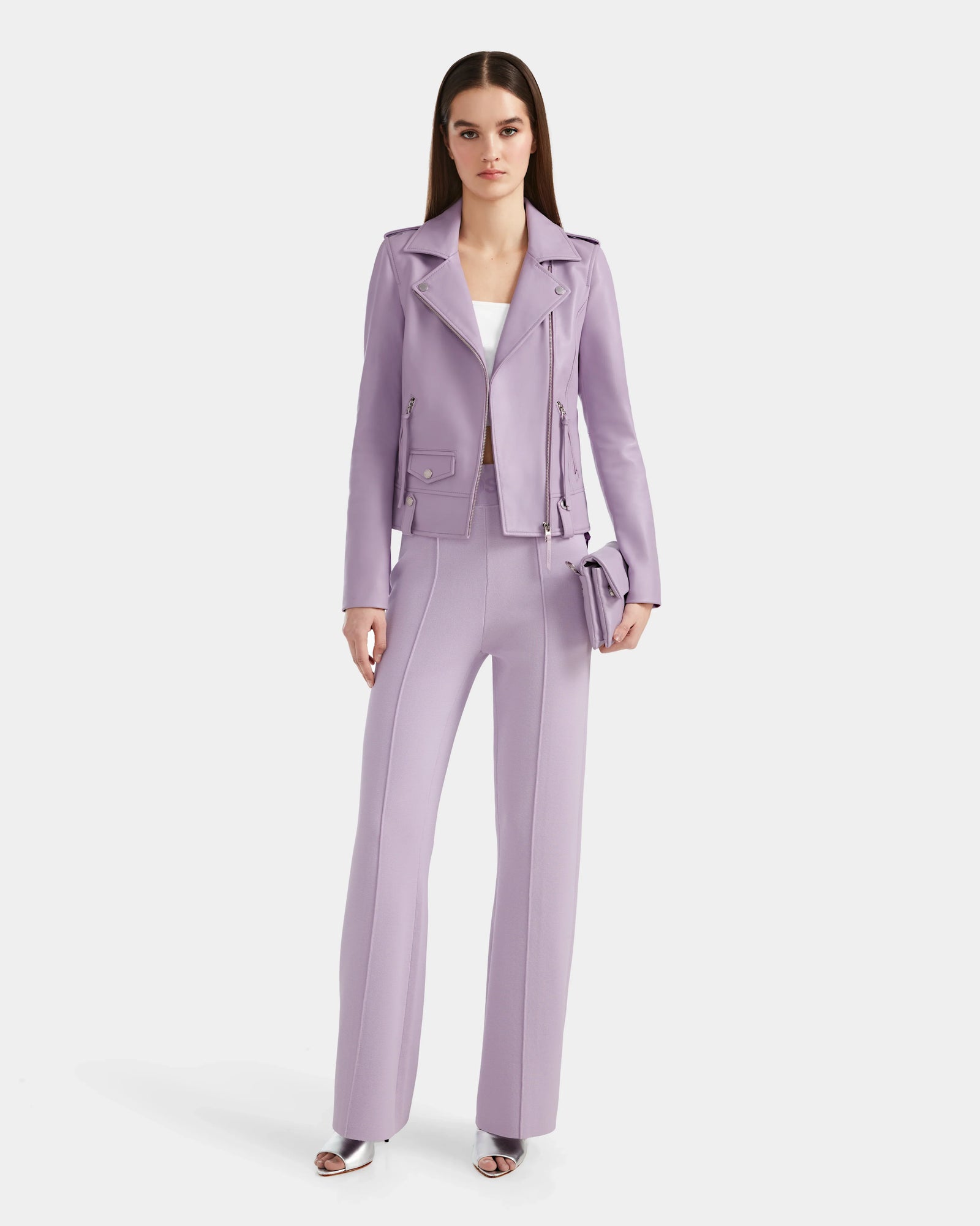 A woman stands against a white background wearing the Rudsak MERGO - 8124034 Lilac Foam pantsuit with flared pants, a white top, silver open-toe heels, and a matching clutch, topped with a Rudsak biker jacket for an edgy touch.