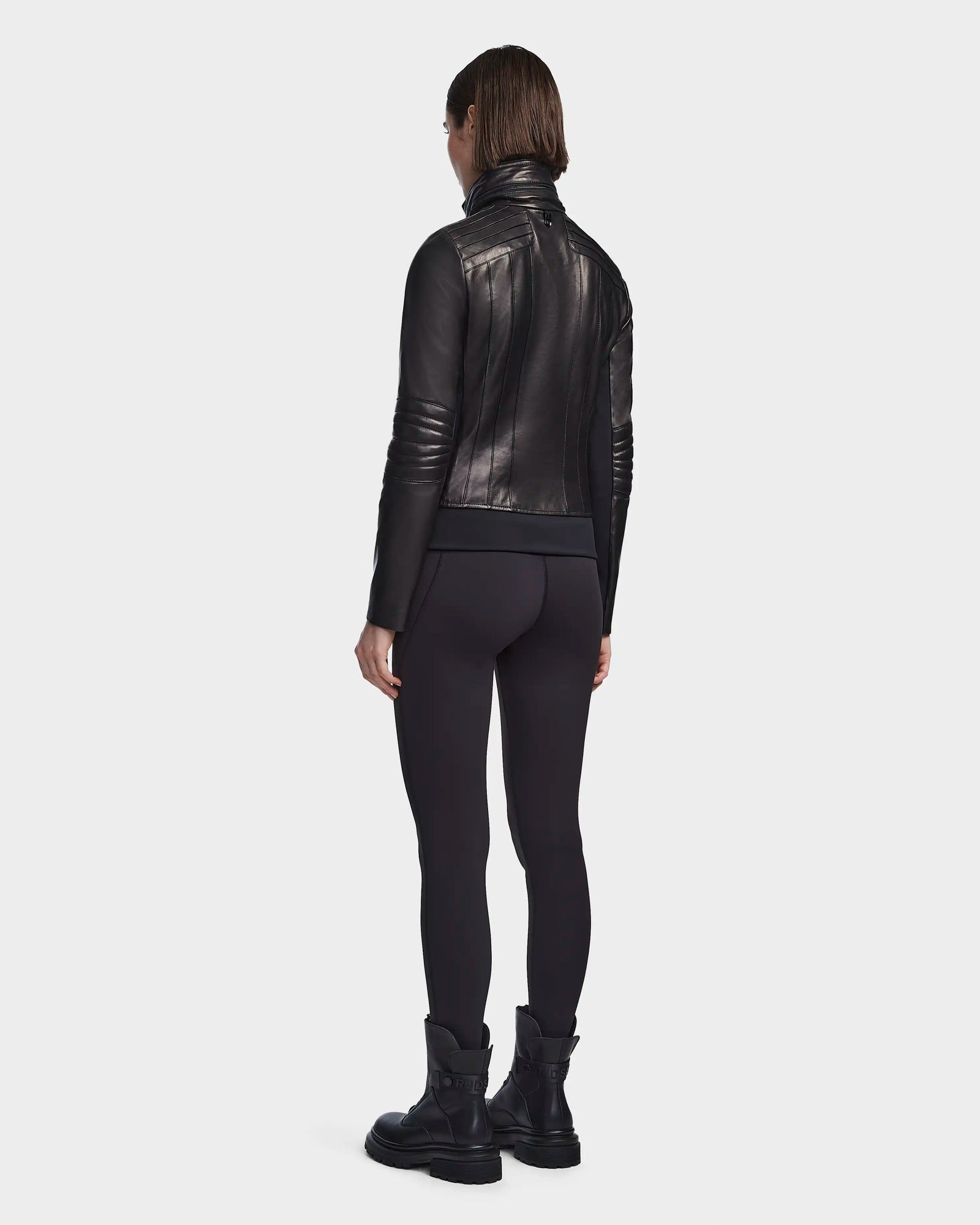 A woman with chin-length brown hair wears the Rudsak PRESLEY - 8122027 BLACK leather jacket, paired with black leggings and boots, standing with her back to a plain light background.