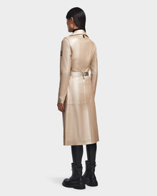 A person with long dark hair stands facing away, wearing the Rudsak KIRALY - 8123553 Metallic Ivory trench coat, paired with black leggings and studded boots against a plain light background.