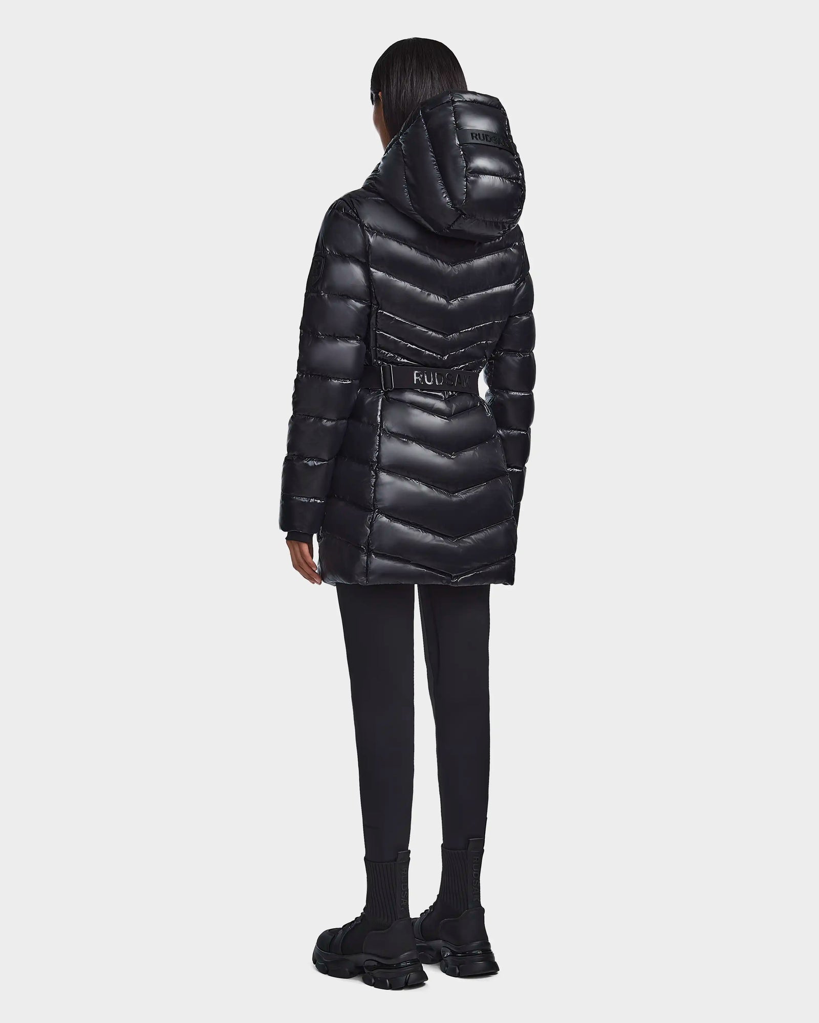 A person wearing the Rudsak MALAYA - L123510 Black women’s down puffer jacket stands with their back to the camera, paired with black pants and shoes, against a plain white background.