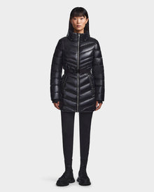 A woman faces forward wearing the Rudsak MALAYA - L123510 Black recycled glossy black women’s down puffer jacket with a hood, paired with black pants and shoes, set against a plain white background.