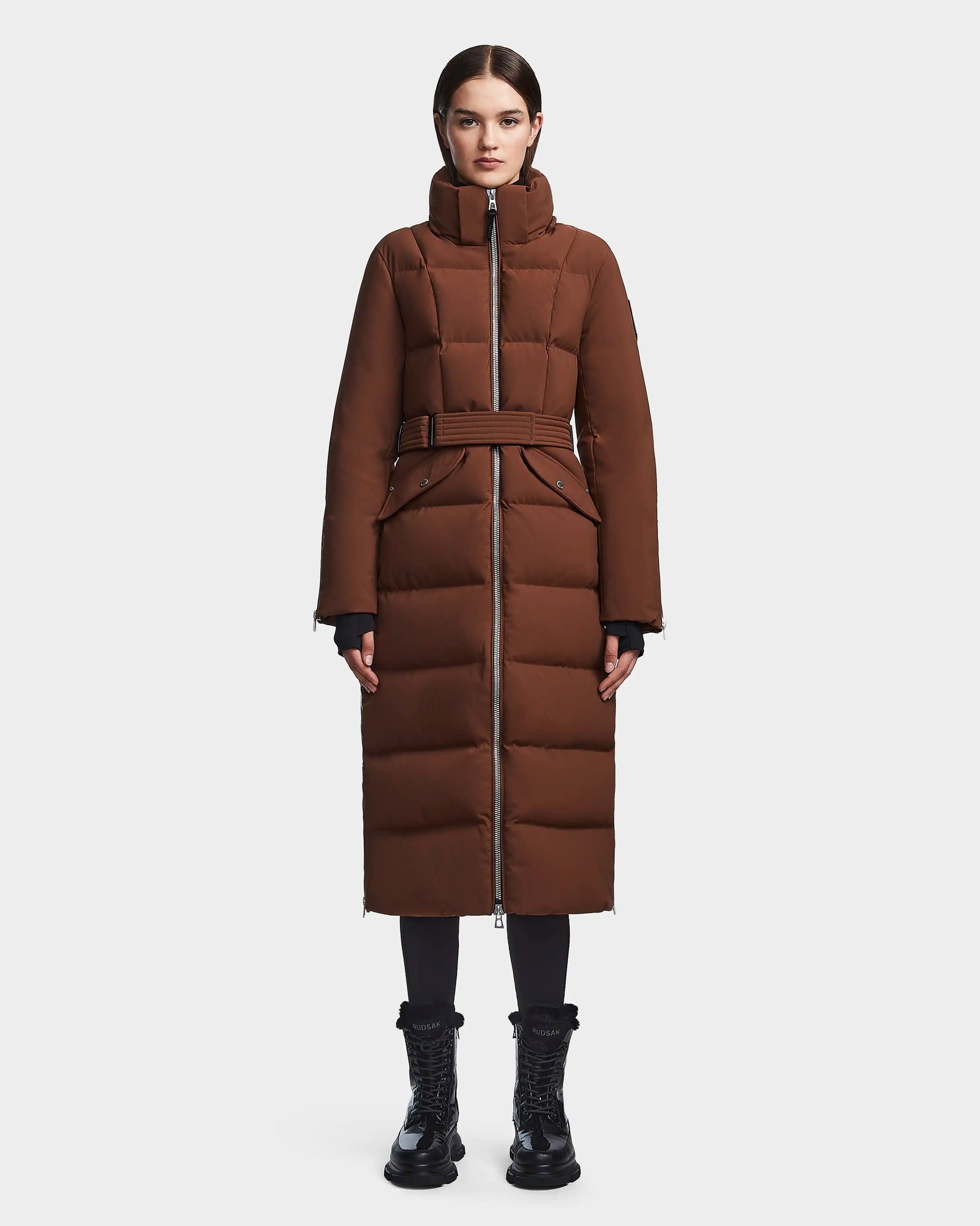 A woman faces forward, wearing the RUDSAK CALI - 8122502 COHIBA long brown RDS-certified down puffer coat with a high collar, black pants, and black lace-up boots against a plain white background.