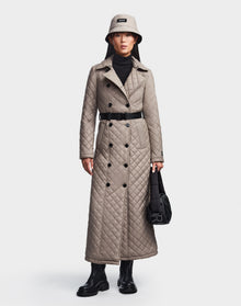 A person wears the Rudsak DAISY - 8123580 Cement women's quilted maxi trench coat with black buttons, styled with a beige bucket hat, black turtleneck, boots, and black handbag against a plain white background.