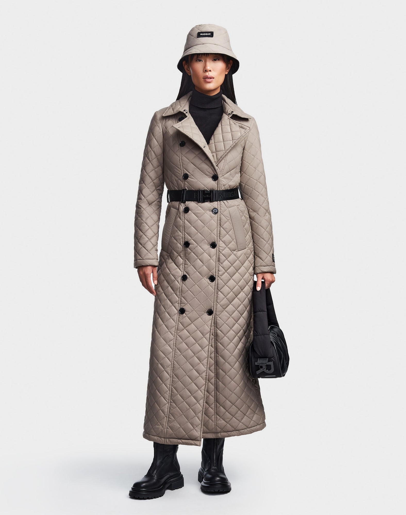 A person wears the Rudsak DAISY - 8123580 Cement women's quilted maxi trench coat with black buttons, styled with a beige bucket hat, black turtleneck, boots, and black handbag against a plain white background.