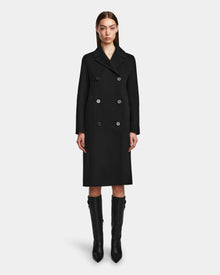 A woman with straight brown hair wears the Rudsak LAYLAH - 8124699 Black knee-length double-breasted women’s wool coat and knee-high black boots, standing against a plain white background.