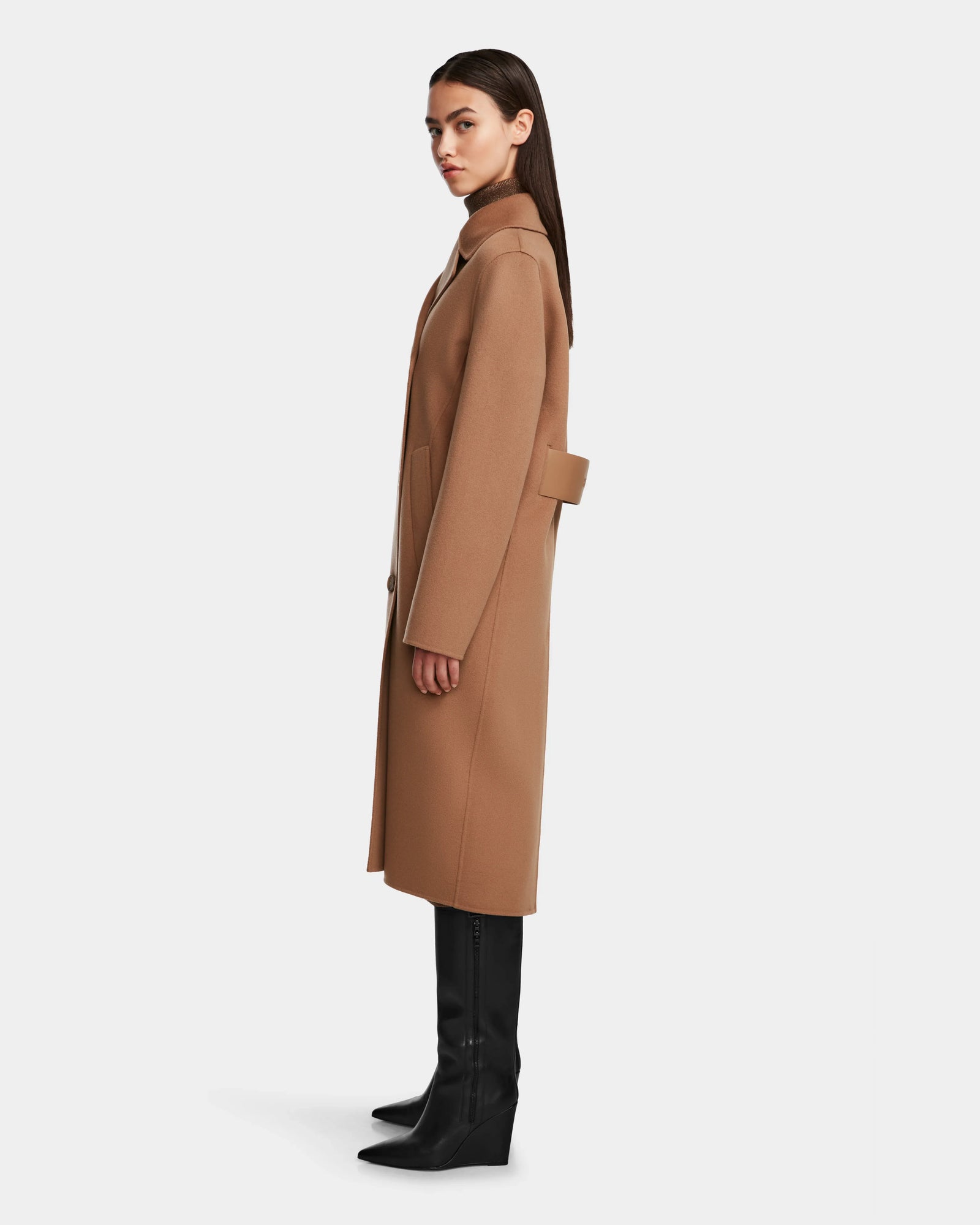 A woman stands in profile wearing the Rudsak LAYLAH - 8124699 Camel knee-length womens wool coat with a back belt detail, black knee-high boots, and straight brown hair against a plain white background.