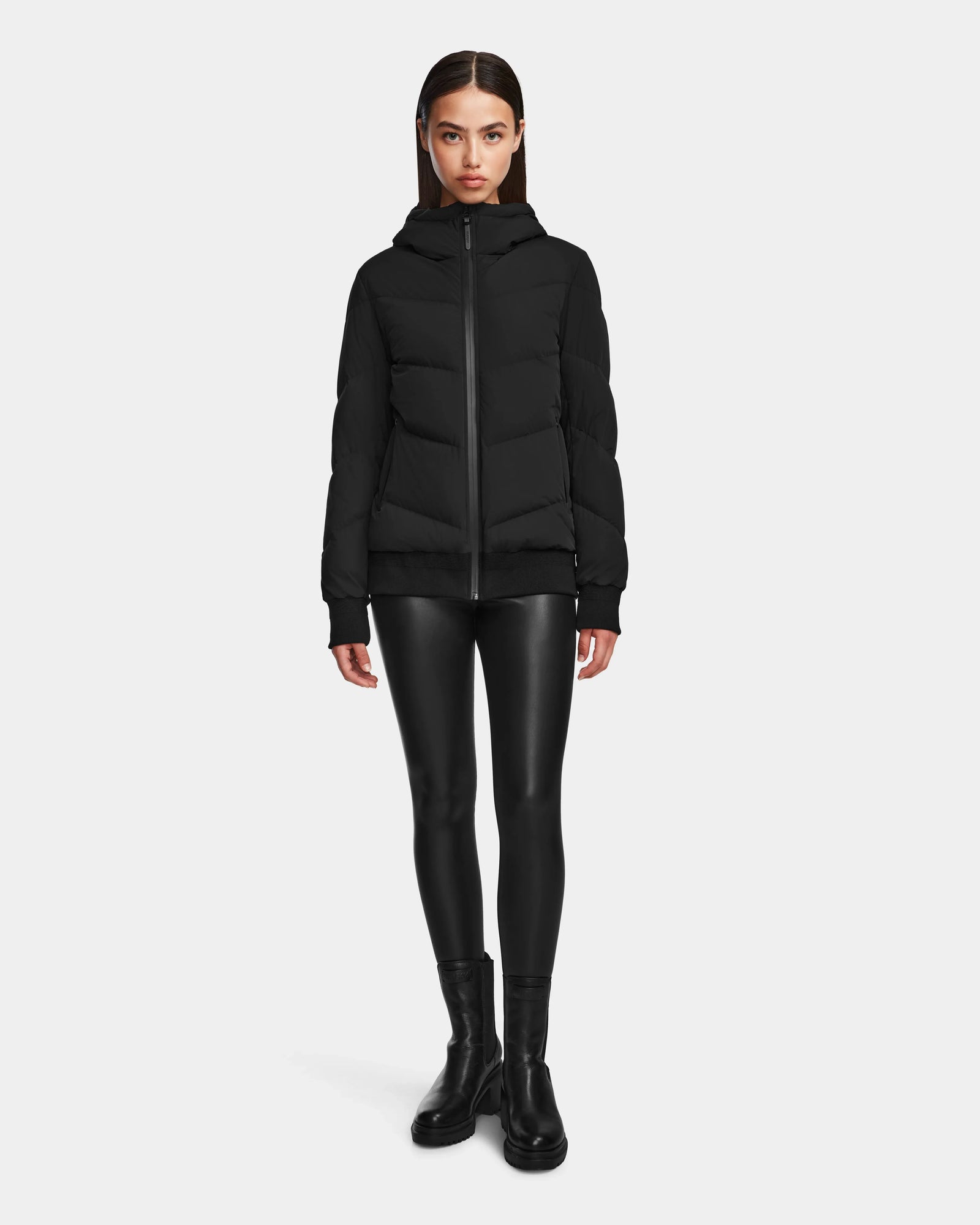 A woman stands against a plain background wearing the Rudsak ARLA - 8124579 Black puffer jacket with leather sleeves, paired with black shiny leggings and ankle boots, facing forward with her arms relaxed by her sides.