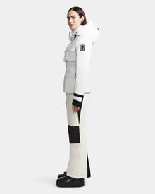 A woman stands in profile wearing the Rudsak LUX - 8123618 Iceland white hooded women’s ski jacket with black accents and flap pockets, paired with matching pants and black boots. The waterproof jacket combines stylish design with protective performance.