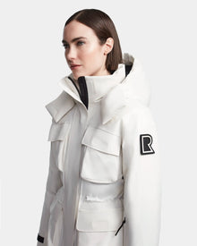 A woman with straight brown hair models the Rudsak LUX - 8123618 Iceland women’s ski jacket in white, with big front pockets, a black R patch on the upper sleeve, and RECCO tech, standing against a plain light background.