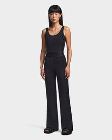A woman stands facing forward in the Rudsak SEDONA - 8523541 Black tank top and flare leg pants, paired with black slide sandals. Her straight black hair with bangs completes her casual chic look against a plain white background.