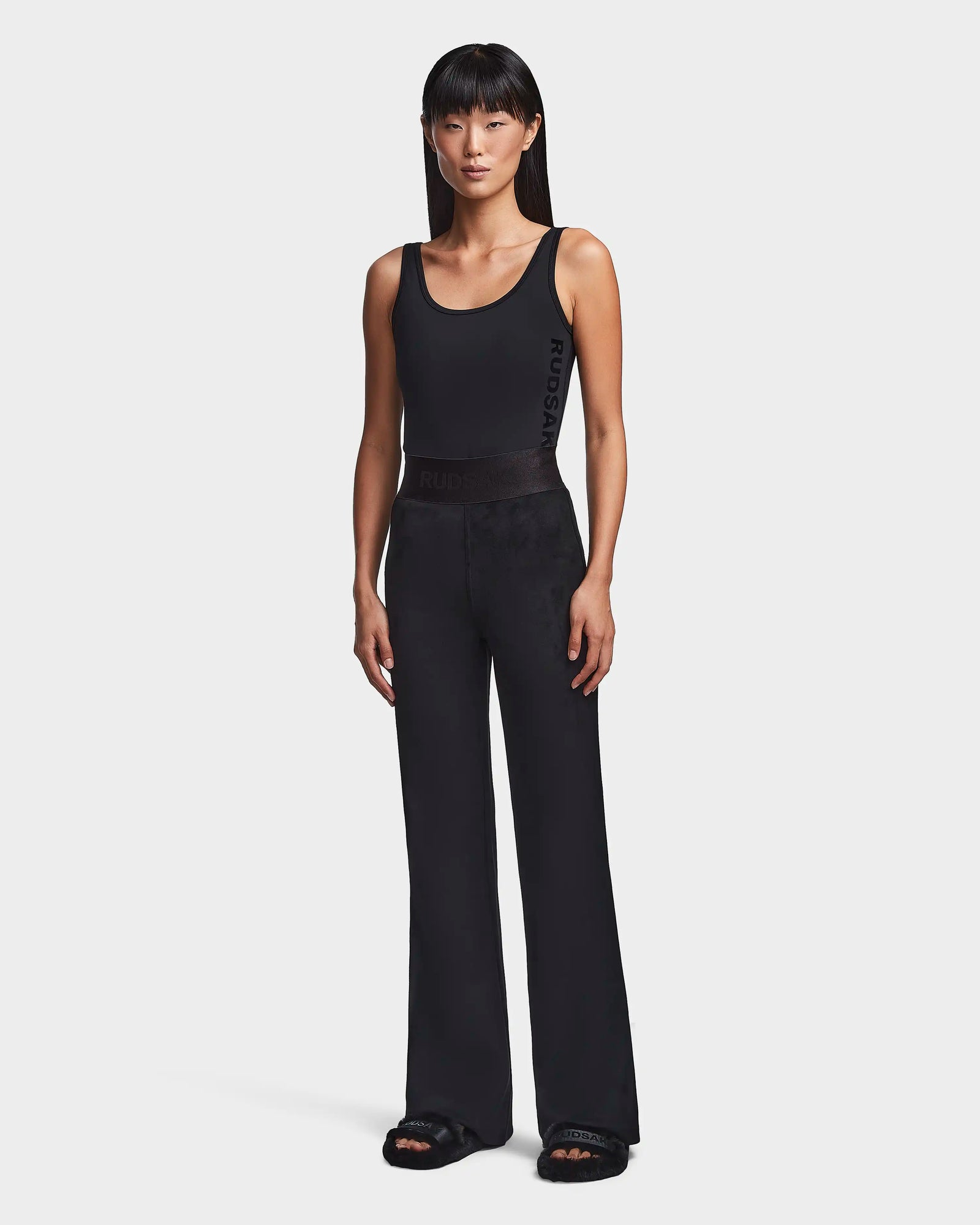 A woman stands facing forward in the Rudsak SEDONA - 8523541 Black tank top and flare leg pants, paired with black slide sandals. Her straight black hair with bangs completes her casual chic look against a plain white background.