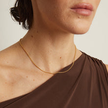 Model. Woman wearing a brown one-shoulder dress with a gold necklace against a neutral background