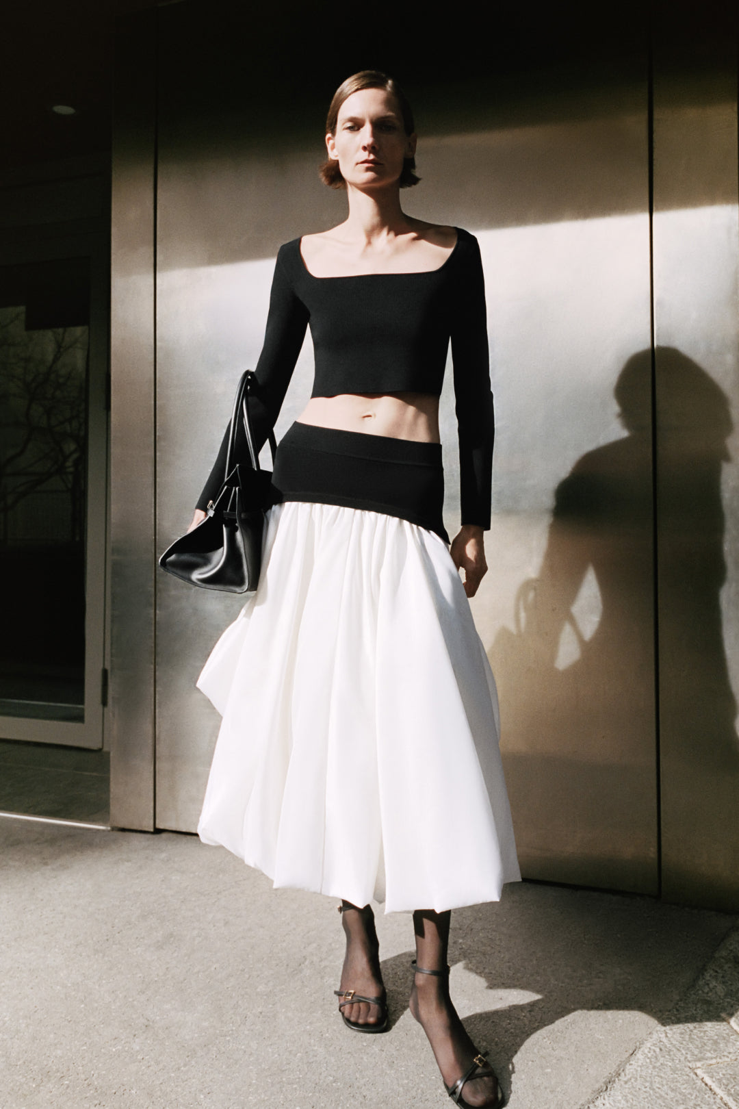 Black cropped knit top with long sleeves and square neckline worn with white midi skirt.