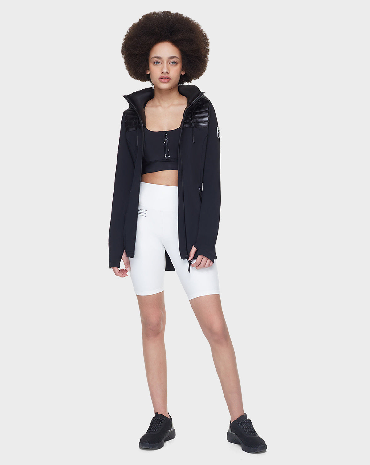 A woman with an afro faces forward, wearing the CAMIRA - 8122011 BLACK sports bra by Rudsak, an activewear jacket, white high-waisted bike shorts, and black sneakers against a plain light background.