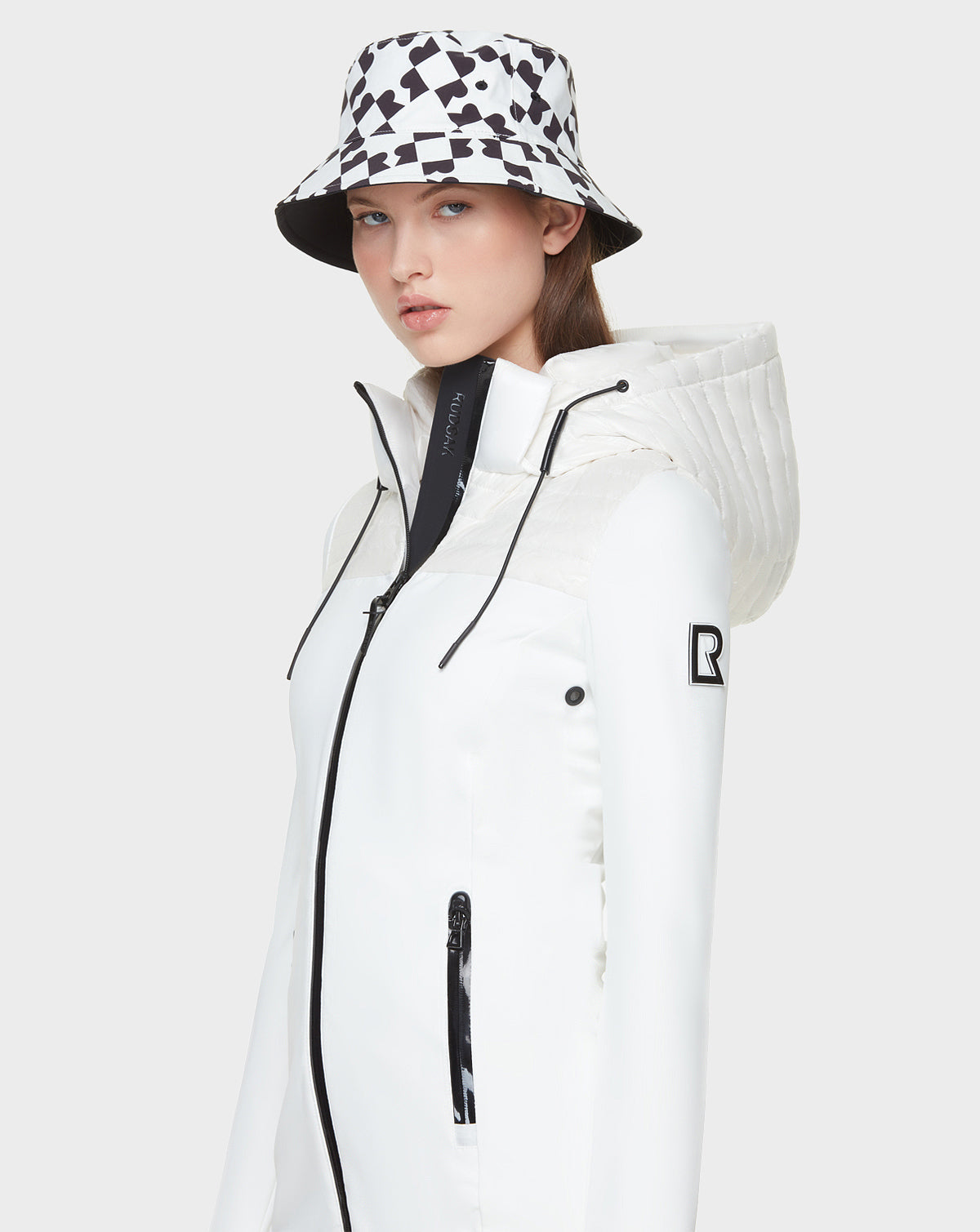 A woman wears the Rudsak CAMIRA - 8122011 WHITE womens activewear jacket with black zipper details and a black-and-white geometric bucket hat, standing against a plain light gray background—stylish comfort for outdoor workouts.